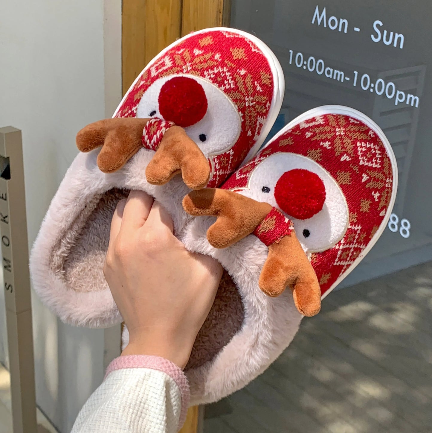 Plush slippers with reindeer design held by a hand, with a store entrance in the background.