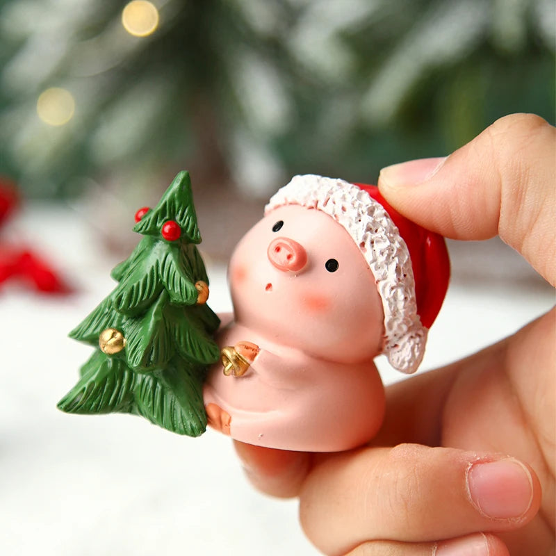 Small pink pig figurine with a red hat holding a green Christmas tree ornament, held by a hand against a blurred festive background.